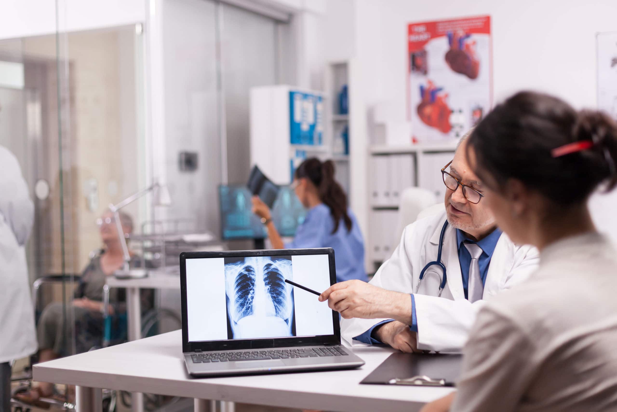 Mature doctor pointing at patient lungs ct scan on laptop during medical visit in hospital office. Medic in white coat discussing with disabled senior woman in wheelchair and nurse in holding x-ray.